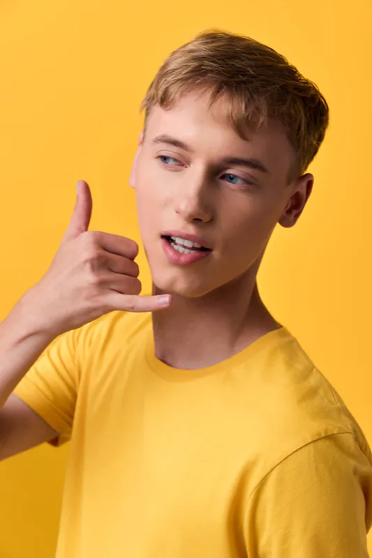 Male youth in a yellow shirt demonstrating a shaka hand gesture against a vibrant yellow background, casual pose, contemporary lifestyle cue for friendly expression, social vibe and approachable