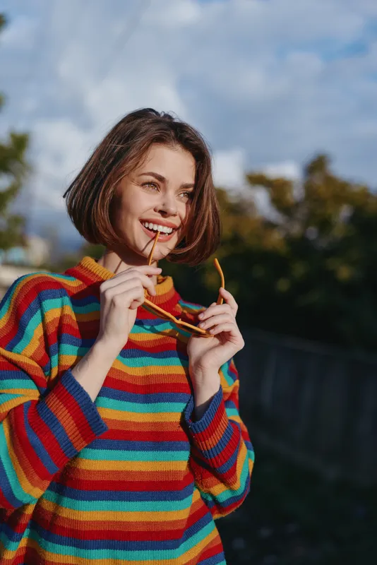 Woman smiling striped sweater sunglasses outdoors short hair casual lifestyle: mid twenties model with bob haircut holding glasses near her mouth, colorful knit portrait in natural light, playful and
