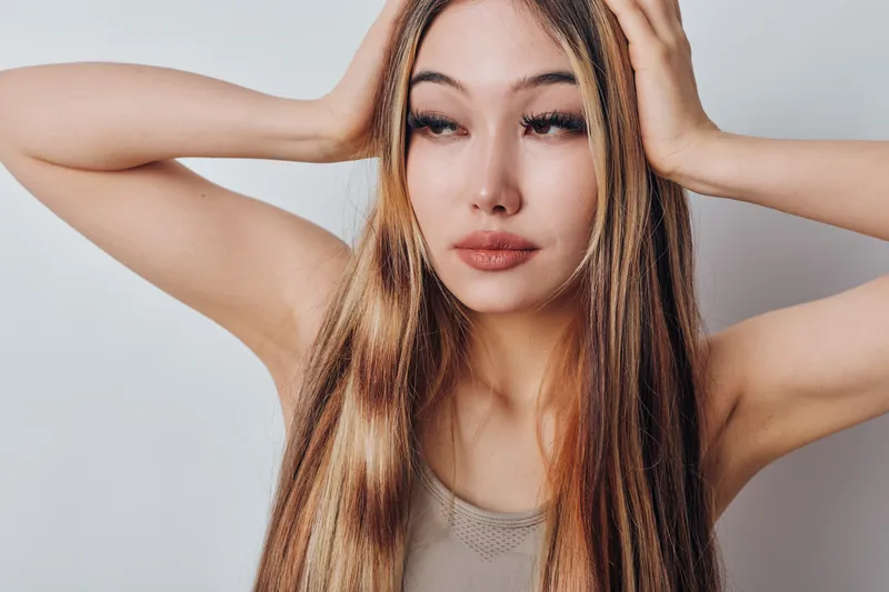 Young woman with long straight hair holding head with hands against white background. Female model with natural makeup posing with closed eyes and neutral expression in studio.