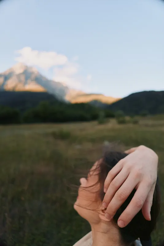 A contemplative woman outdoors in a sunlit field with distant mountains, hand on her head, expressing reflection, calm, and personal growth in a natural setting.