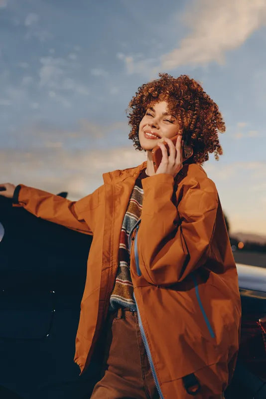 Woman lifestyle outdoors sunset curly jacket portrait smiling stands by car at dusk holding phone near face calm confident gesture wearing orange coat scarf