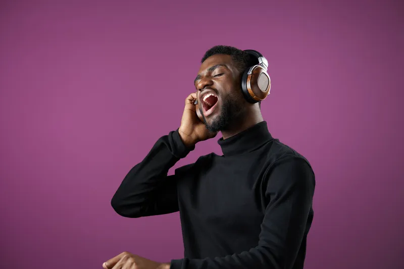 Young Black man singing joyfully with headphones against a vibrant purple background, showcasing happiness and passion for music