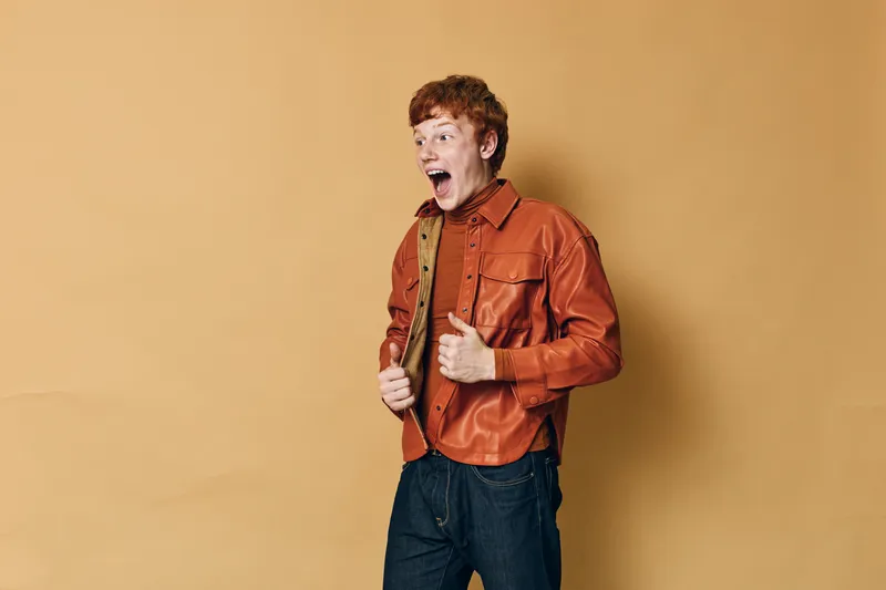 Young man with red hair in orange jacket and jeans expressing surprise or shock with open mouth against beige background. Casual clothing and emotional reaction.