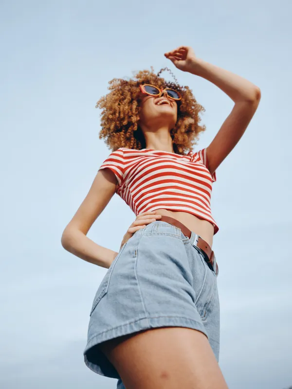Vibrant portrait of a carefree young woman in a striped crop top and denim shorts, standing confidently under a bright blue sky with warm sunlight.
