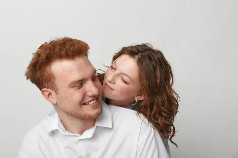 Smiling young couple with red hair and curly brown hair share a tender moment, looking at each other with affection on a plain white background. Concept of love, intimacy, and happiness.