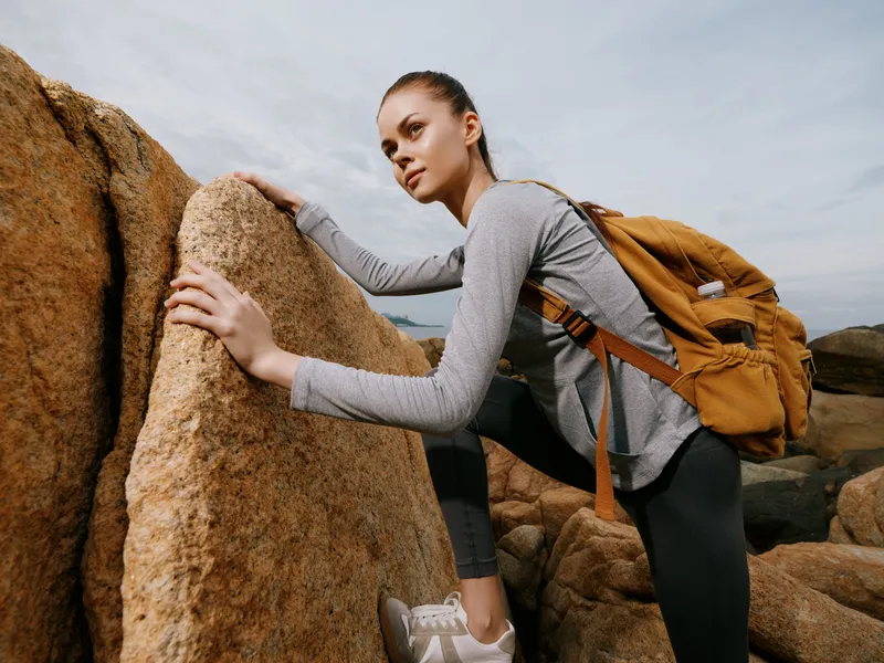 Stylish female hiker climbing rocky terrain with a backpack, showcasing determination and adventure, with a natural landscape background