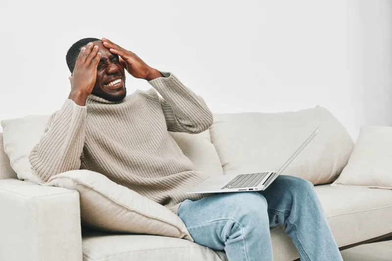 A distressed young man in a cozy sweater expressing frustration while working on his laptop at home