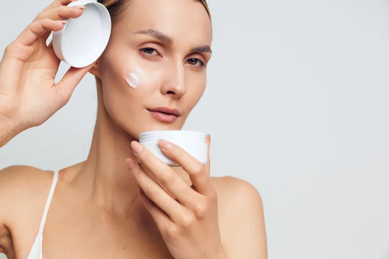 Young woman applies moisturizing cream on face holds open jar with white lotion looks at camera with natural skin isolated on light background skincare beauty concept.