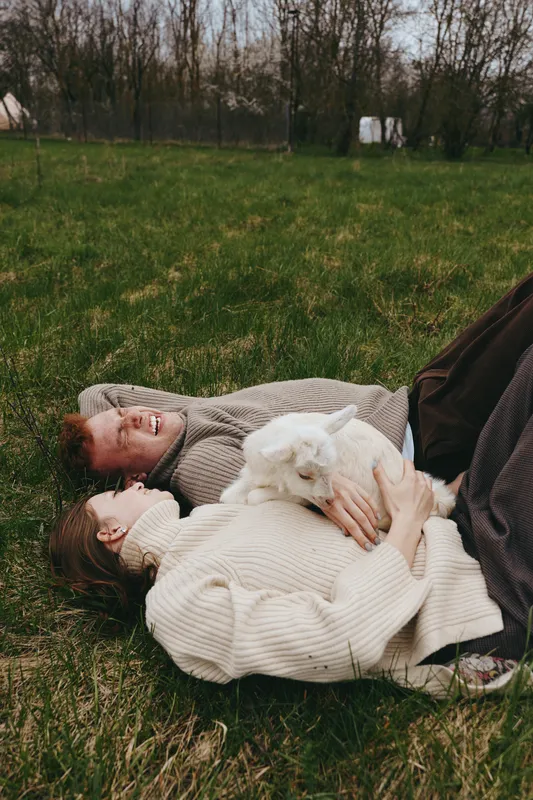 Lifestyle outdoor scene with a couple lying on grass, cuddling a small goat kid, soft sweaters, relaxed smiles, natural light, tranquil countryside mood, close connection, gentle warmth, calm