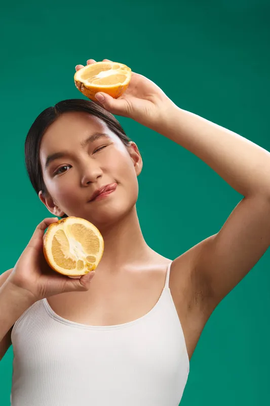 Asian woman holds lemon halves above her head against a green backdrop, exuding brightness and confidence with a playful pose and fresh vibe.