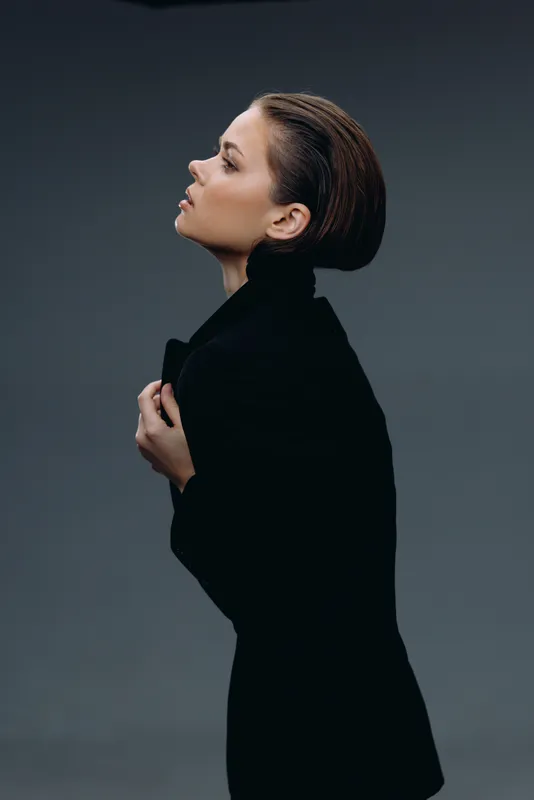 Profile of a stylish woman wearing a black coat, posing thoughtfully against a dark gray background.