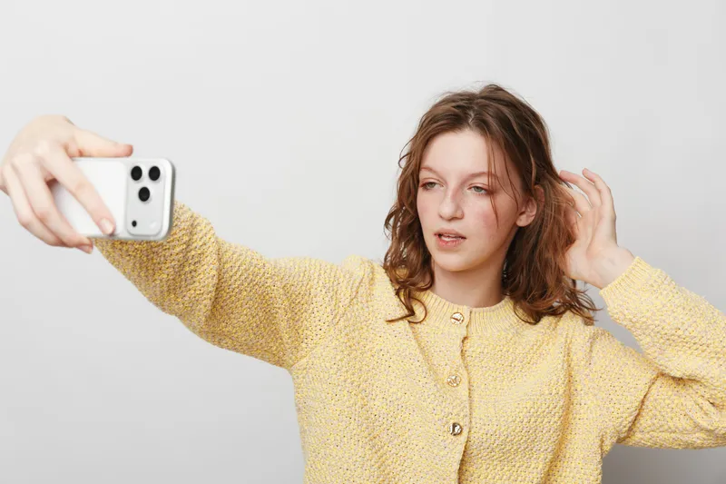 Young woman in yellow sweater takes selfie with smartphone, looking at screen with relaxed expression. Indoor casual portrait on plain light background, concept of social media and communication.