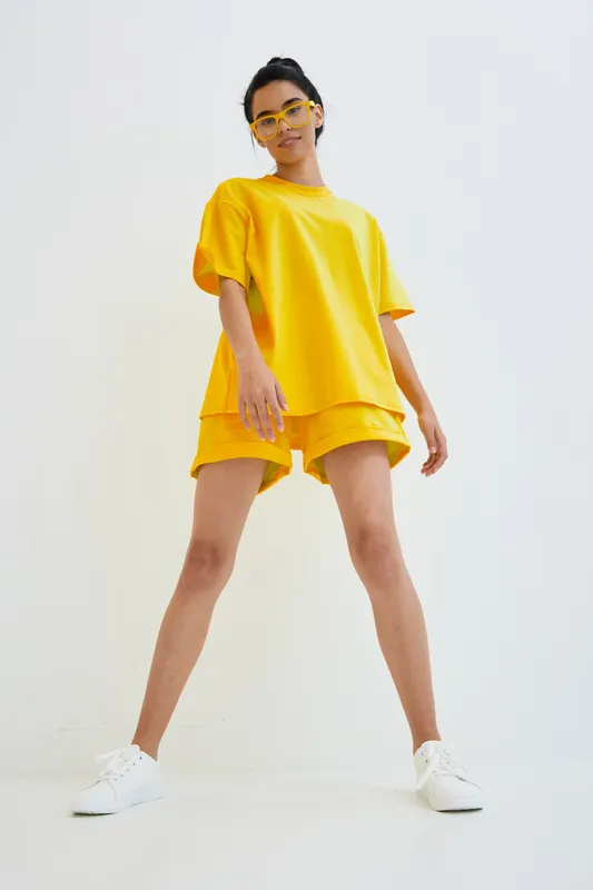 Young woman wearing bright yellow casual outfit and glasses stands confidently against a white background, showcasing modern street style and summer fashion.