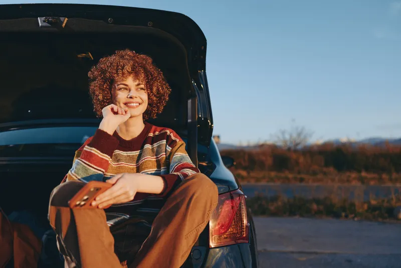 Smiling woman with curly hair sits on car trunk by the road at sunset, casual autumn sweater, outdoor lifestyle scene, warm evening light, relaxed travel mood