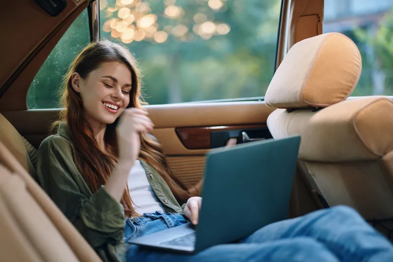Young woman enjoying her time working on a laptop in a car, with a joyful expression and a natural, relaxed outfit, surrounded by greenery.