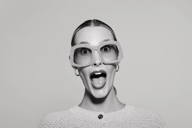 Surprised young white woman with fair skin and slicked hair wearing large glasses and a sweater. Minimalist studio portrait in black and white with open mouth expression.