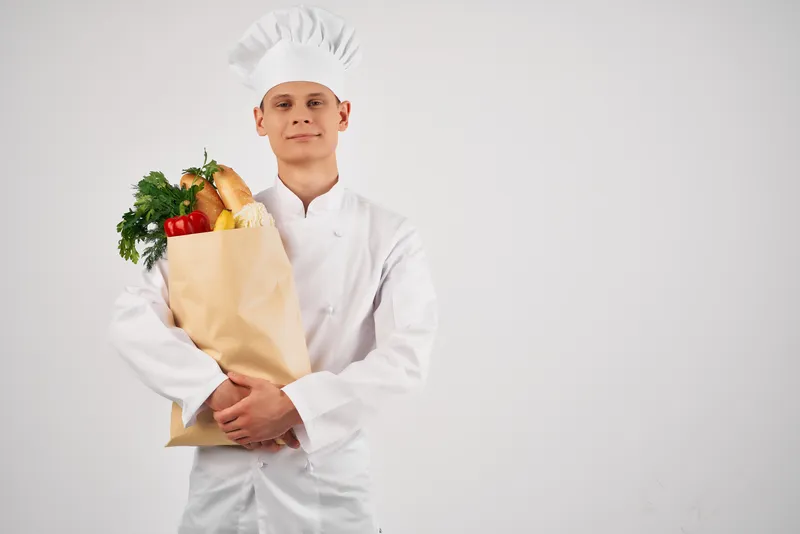 Мужчина в униформе шеф-повара с пакетом продуктов жестикулирует рукой, здоровая еда на кухне