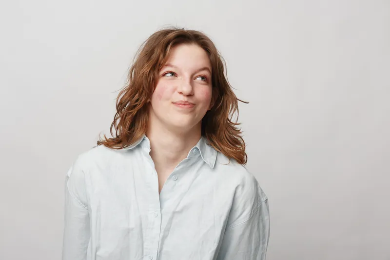 Young woman with curly hair wearing white shirt looks up with playful smile on light background. Concept of casual style, positive emotions, and natural beauty in studio portrait.