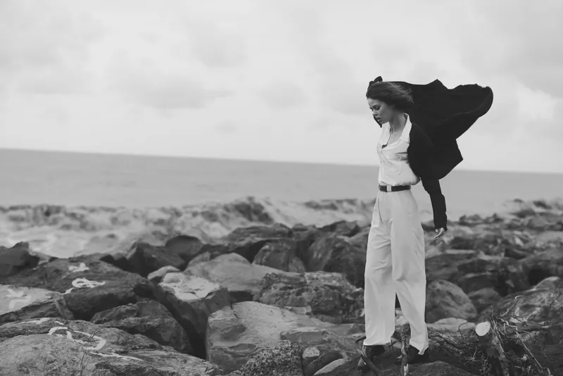 Wind swept figure on a rocky shoreline wearing white suit and flowing cape, creating a dramatic monochrome fashion moment by the sea and rugged coast lines