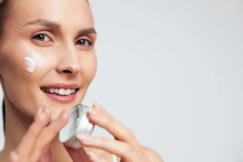 A smiling woman applies white cream on her cheek using fingers, holding an open jar in a bright studio. Concept for skincare, beauty routine, and healthy skin care.