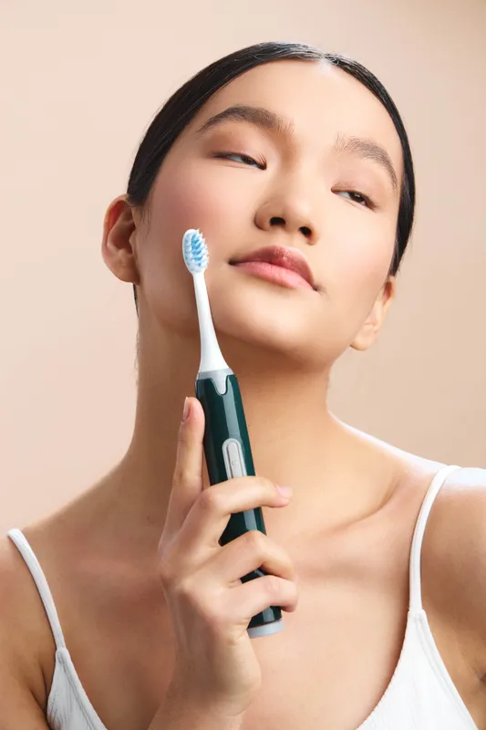 Asian woman holds a toothbrush near her face in a clean, softly lit setting, smiling with calm confidence as she demonstrates daily personal care and hygiene.