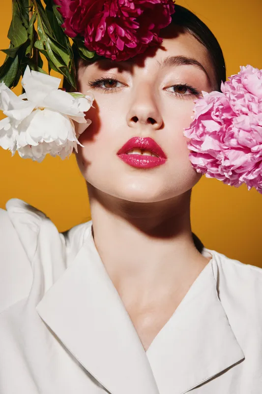 Close-up portrait of a young woman with glossy red lips holding pink and white flowers against a bright orange background. Creative fashion and beauty concept.