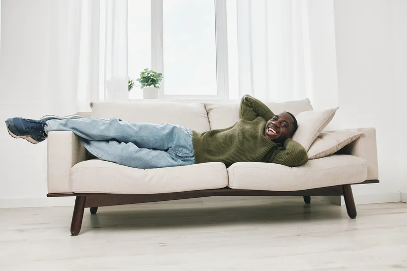 Happy Black man relaxing on a modern sofa in a cozy white living room, wearing casual green sweater and blue jeans, showcasing comfort and joy