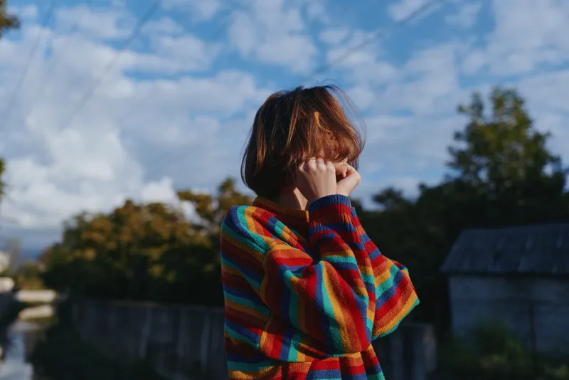 Person in rainbow sweater covering face outdoors in a candid portrait, shy pose and colorful knit against cloudy sky and backyard trees for lifestyle and emotion.