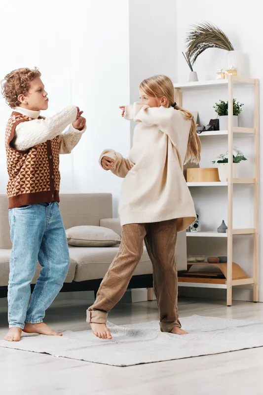 Two children engage in playful roughhousing indoors, tossing cushions and sparring softly in a cozy living room with light decor and a cheerful, energetic atmosphere.