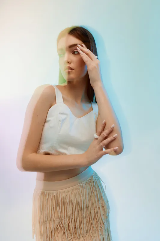 Young woman in a white top and beige skirt poses with a thoughtful expression. Soft lighting and subtle color overlays create a fashion and beauty concept in studio.