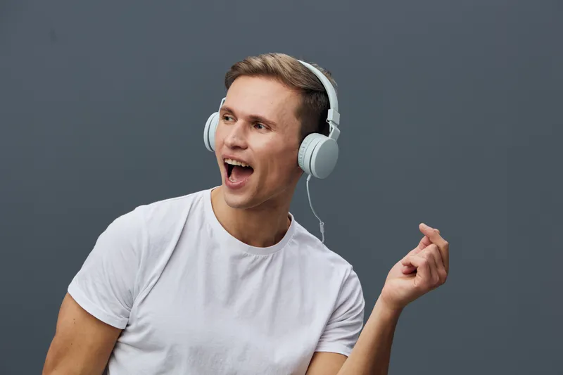 Happy young Caucasian man wearing white t-shirt and headphones enjoying music, smiling and singing with closed eyes, on plain grey background. People lifestyle concept, emotion, relaxation, audio, ent