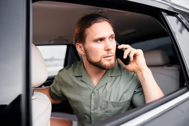 Бородатый длинноволосый мужчина сидит в автомобиле и разговаривает по телефону, демонстрируя сосредоточенность