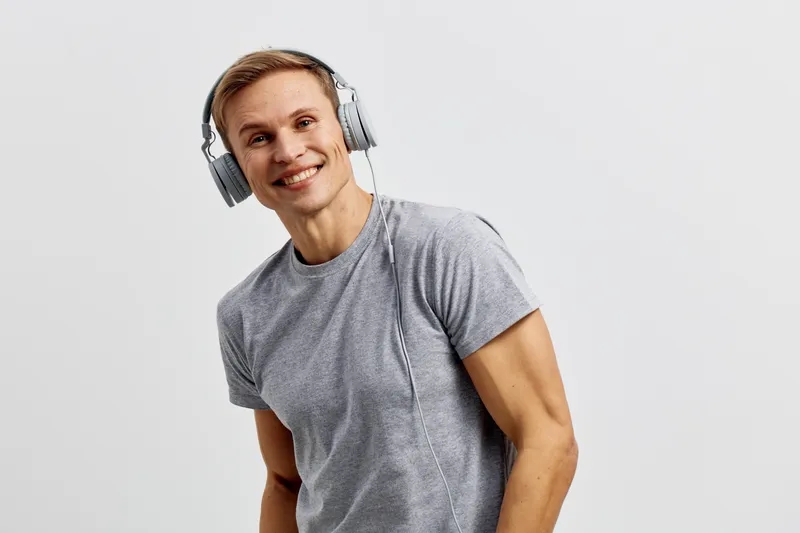 Smiling young man with short blond hair wearing gray t-shirt listening to headphones and looking at camera, casual attire, upbeat mood, indoor studio shot. People lifestyle concept