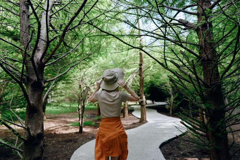 Woman hat path forest trees nature walking trail captured from back view on a winding route, casual summer outfit with orange skirt, peaceful outdoor exploration amid lush greenery.