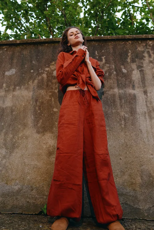 Cheerful woman outdoors, confident pose in rust outfit, sunlit moment, natural colors, leafy backdrop, casual elegance, relaxed lifestyle