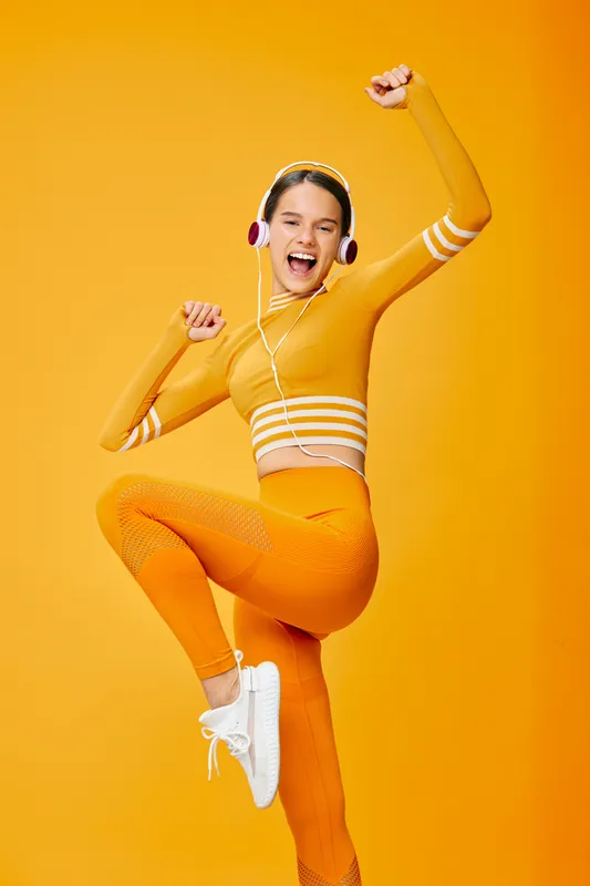 Fitness woman in athletic orange outfit jumps with headphones in a bright studio, conveying energetic movement and athletic confidence during a high-energy workout session.
