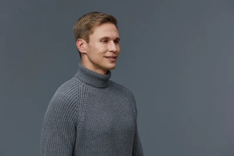 Close-up of a smiling young man with blond hair wearing a gray turtleneck sweater, looking to the side, casual indoor setting, clear gray background, studio portrait, lifestyle concept, cheerful and r