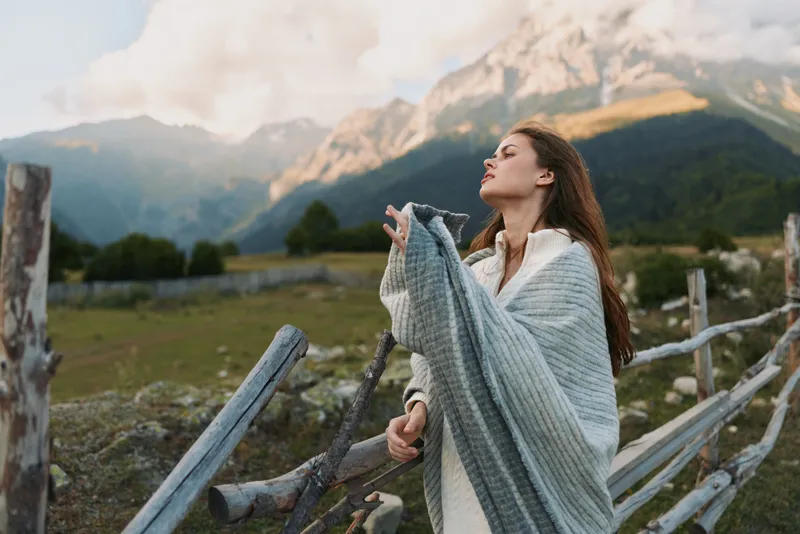 Woman outdoors, embracing nature in a mountainous landscape, wrapped in a cozy shawl, eyes closed, enjoying a breezy moment and warm light.