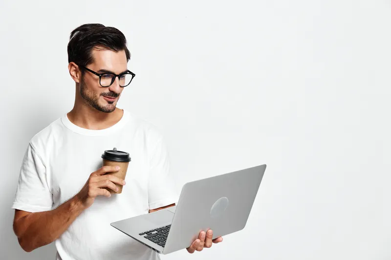 Happy smiling young man holding a coffee cup while using a laptop computer, wearing casual white t-shirt, standing against plain solid light background.