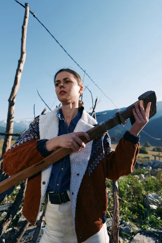 Woman outdoors in rural mountains holding a wooden staff, portrait of a young farmer in layered jacket and shirt with braids, rustic fence and natural landscape in sunlight.