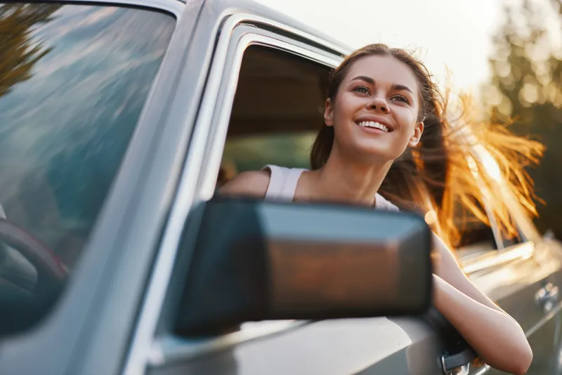 Happy young woman enjoying a road trip with wind in her hair, smiling while leaning out of a car window, embodying freedom and adventure.
