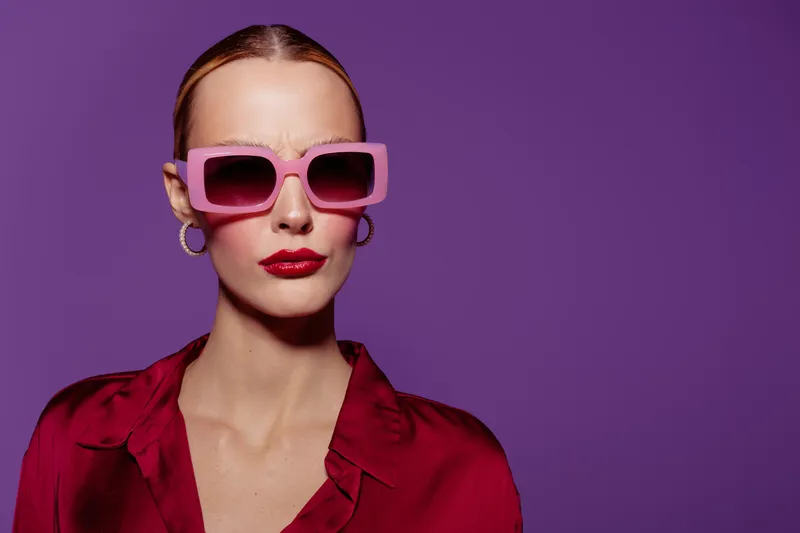 Stylish woman with slicked hair wears pink sunglasses and red blouse against purple background. Fashion portrait with bold makeup and confident expression in studio lighting.