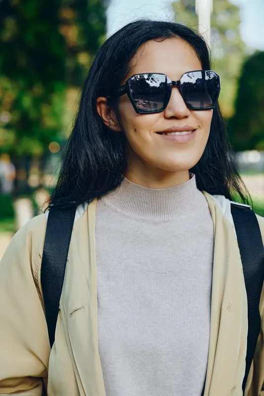 Woman wearing sunglasses, beige coat and backpack outdoors, smiling in a park, casual street style, natural lighting, candid lifestyle shot for fashion, travel, or everyday moments