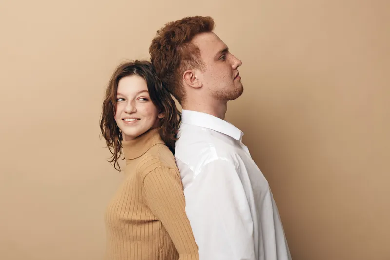 Young man and woman stand back to back against beige background. The woman smiles looking sideways while the man looks forward with a neutral expression. Concept of contrast and relationship.
