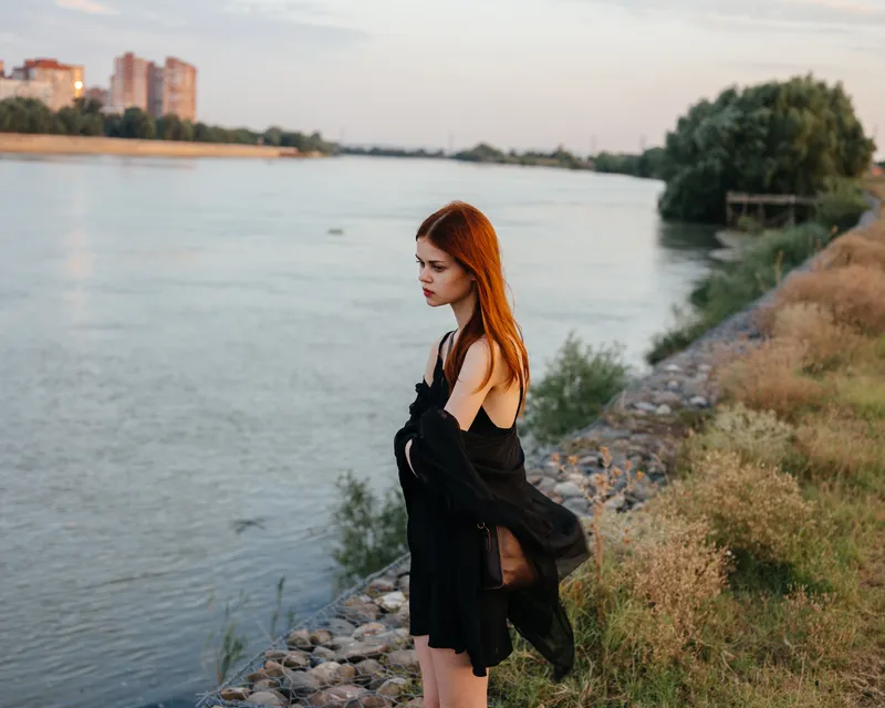Рыжеволосая женщина в черном платье стоит у реки на природе