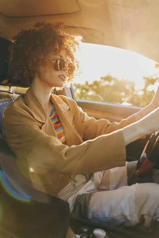 Young woman with curly hair drives a vintage car at golden hour, wearing a tan jacket and striped shirt, smiling softly as sunlight fills the cabin.