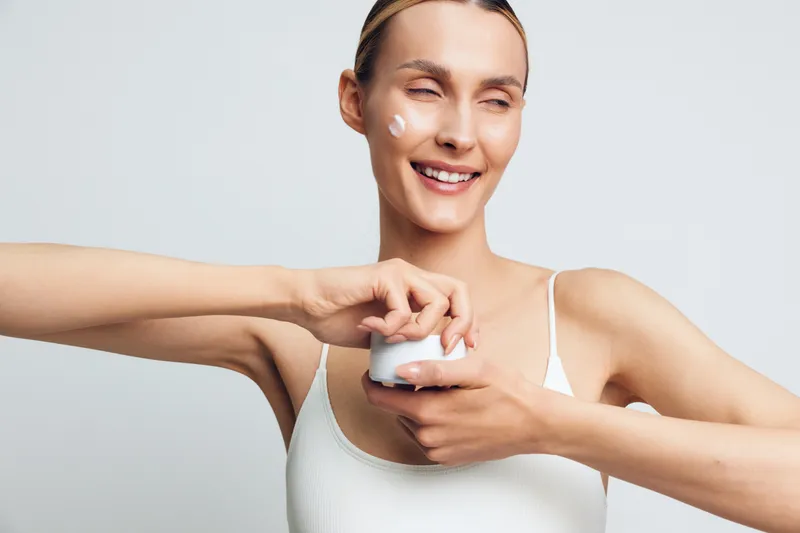 Woman skincare cream face moisturizer hygiene care beauty spa wellness applying lotion in bright studio, smiling with relaxed posture, minimal gray background, hygienic self routine