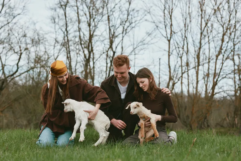 Three young adults enjoy a spring day in the countryside playing with two baby goats on green grass surrounded by bare trees, capturing a joyful lifestyle moment with casual clothing.