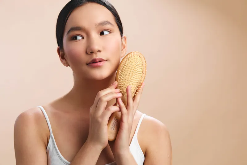 Asian woman holds a brush up to her hair with a calm expression in a soft studio setting, conveying beauty, self care, and focused personal grooming, and professional presence for a modern lifestyle