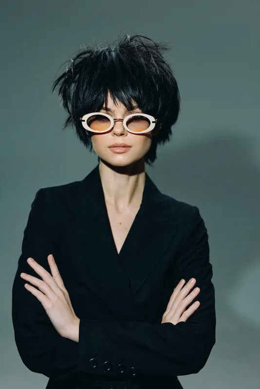 Stylish woman wearing round sunglasses and black blazer with crossed arms against plain background.